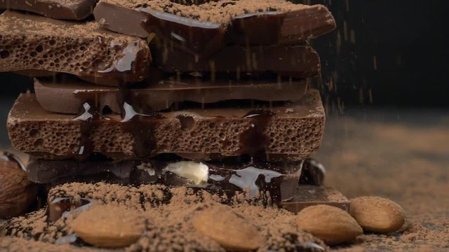 Cocoa powder falling onto heap of various chocolate pieces and nuts. Slow motion, slider shot