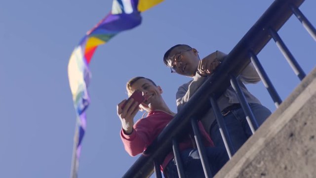 Happy Gay Couple Stand Under Gay Pride Flag And Take Selfies Together