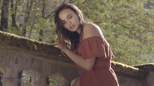 Dreamy Bohemian Model, Bathed In Golden Sunlight, Poses Against Stone Bridge In Enchanted Forest 