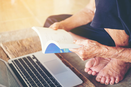 Old senior woman open read a book on hand background notebook labtop on wooden, Thailand