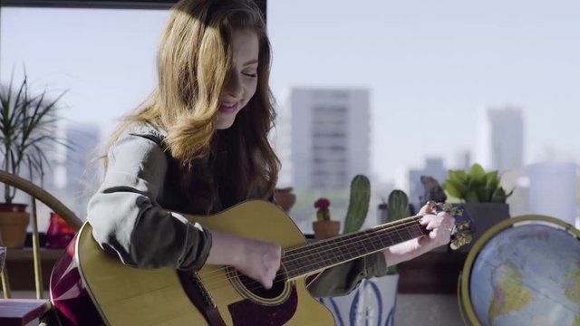 Happy Young Woman Practices Playing Acoustic Guitar In Her Trendy City Apartment