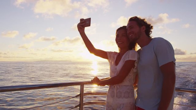 Smart phone selfie - Romantic couple taking selfie by sunset over the ocean on small cruise ship sailing on open sea. Woman and man taking cell phone photos on boat travel sailing during vacation.