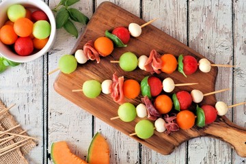 Summer fruit skewers with melon, cheese and prosciutto on a wood serving board, above scene on...