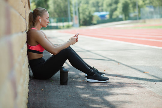 Young sporty woman athlete in sportswear sitting, listen music with headphones and relaxing on stadium track after hard workout