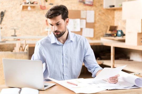 Concentrated youthful bearded lumber craftsman working via gadget at desk
