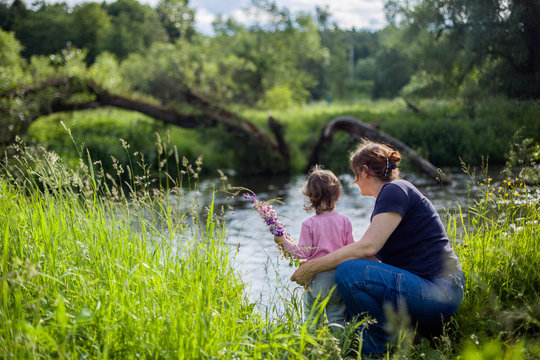 Small toddler girl and and adult woman sitting on the river bank,hugging, watching on water. Summer day, back view, horizontal picture