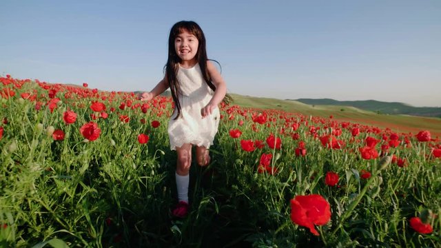 little Asian girl in white dress fun runs in field of red poppies on a Sunny spring day in slow motion