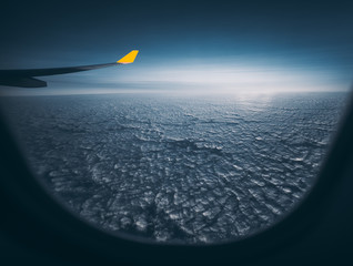 Wide angle shooting through airplane window of aircraft wing with yellow end, even maritime cloudscape and dark blue morning horizon, cirrostratus clouds in distance