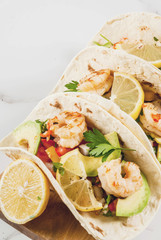 Seafood. Mexican food. Tortilla tacos with traditional homemade salsa salad, parsley, fresh lemon, avocado and grilled shrimp pawns. On a white marble background.