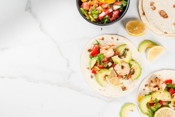 Seafood. Mexican food. Tortilla tacos with traditional homemade salsa salad, parsley, fresh lemon, avocado and grilled shrimp pawns. On a white marble background. Top view copy space
