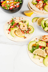 Seafood. Mexican food. Tortilla tacos with traditional homemade salsa salad, parsley, fresh lemon, avocado and grilled shrimp pawns. On a white marble background. Copy space