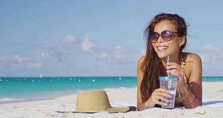 Woman on beach travel vacation smiling and laughing wearing sunglasses relaxing sunbathing lying in sand. Girl on beach vacation in bikini sun tanning relaxing on beach holiday drinking water.