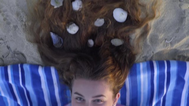 Teen Girl Lays Her Head On A Towel On Beach, Her Hair Has Been Decorated With Seashells, She Looks Like A Mermaid