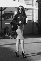 Young beautiful stylish woman wearing green dress, black jeans jacket and sunglasses walking at the street in city. Pretty brunette girl with long hair holding backpack and coffee