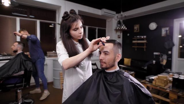 Young female barber making stylish hair to man in salon