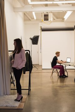 Female model standing by clothes rack with photographer using laptop