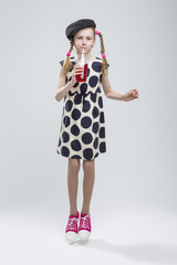 Full Length Portrait of Jumping  Caucasian Girl With Pigtails Posing in Gray Velvel Cap and Polka Dot Dress with Cup of Red Juice. Drinking Through Straw. Against White.