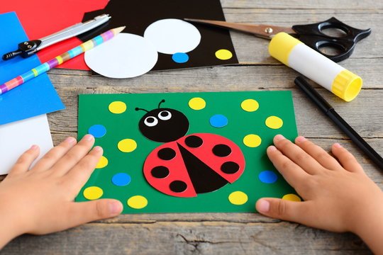 Small child shows a ladybird card. Child made a card with ladybird. Stationery set on a desk. Preschool paper project. Developing children fine motor skills