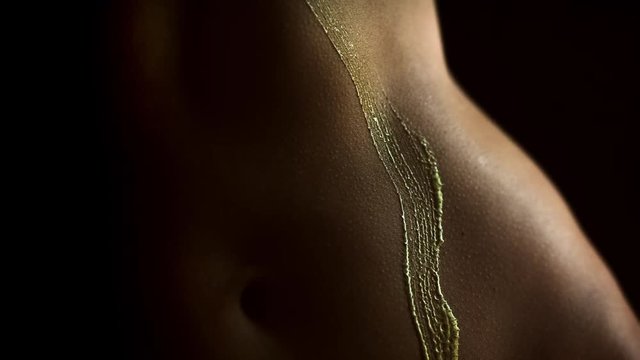Body painting sexual game. Brush with golden color moving up on nude woman body from waist to chest. Studio light, black background