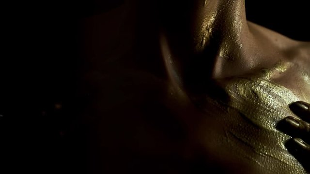 Body painting sexual game. Woman`s hands with golden color caress nude neck, collarbones, moving up and down. Darkness, studio light, black background