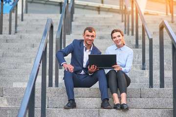 Happy business couple with laptop. Cheerful people sitting on stairs. Web development agency.