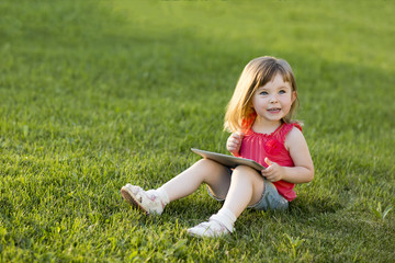 Fototapeta premium Cute little girl is sitting with a tablet on the grass in the park. Emotional portrait. Early education.