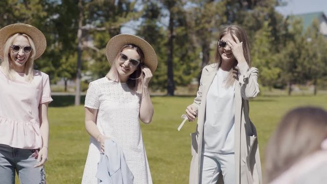 Three beautiful young women in sunglasses embrace, have fun and are photographed against the backdrop of greenery in a summer park. Girlfriends walk and spend time together in the park.