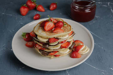 Delicious pancakes on grey table with fruits. Plate full of pancakes with strawberries, bananas and chocolate cream. Berry jam in glass jar