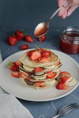 Delicious pancakes on grey table with fruits. Plate full of pancakes with strawberries, bananas and chocolate cream. Pouring over by Syrup. Berry jam in glass jar