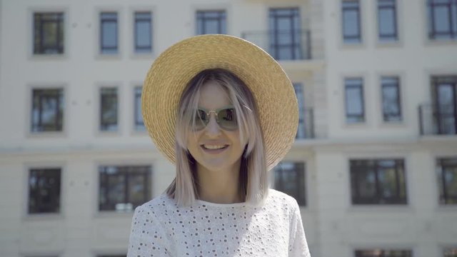 Portrait of a stylish and modern young woman in a straw hat. She stands on the background of a modern building with a white facade, smiles, the wind blows her hair, she sends an air kiss.