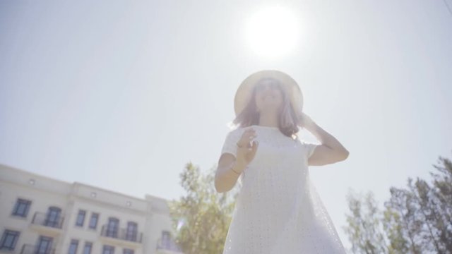Stylish and fashionable girl in a white dress and straw hat stands on the background of a modern building with a white facade, smiling, the wind blows her hair. She is posing and enjoying the weather.