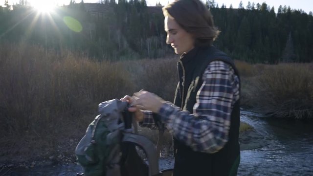 Adventurous Young Man Takes Off His Backpack, Sits Down By Creek, And Searches For Something Inside 