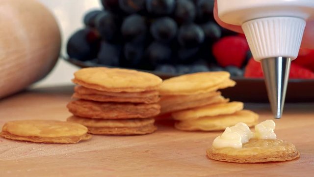 A woman makes a dessert out of phyllo pastry rounds and cream, a plate of fruits in the blurry background - closeup