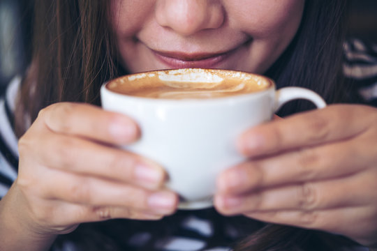 Closeup image of Asian woman smelling and drinking hot coffee in white mug with feeling good in cafe