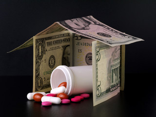 mixed pills and tablets spilled from plastic bottle in house made from banknotes on black background