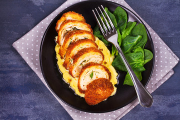 Chicken Kiev on mashed potato with spinach. Breaded chicken breast stuffed with herbs and butter on black plate. View from above, top studio shot