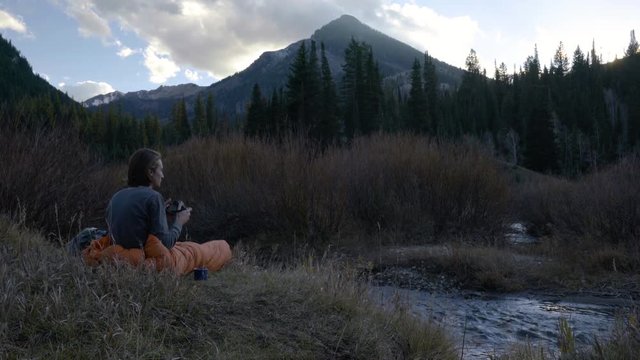 Young Man Sits Inside His Sleeping Bag, And Photographs Mountain Landscape 