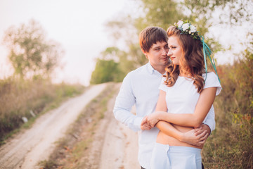 Obraz premium A loving couple are standing in the field road with trees at sunset, holding hands and kissing each other. Bright sunlight shines. Around the greens. Walk on the nature. Country style.