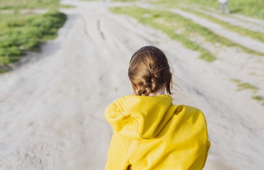 Image of a girl taken from the back in spring. Active healthy child walking alone on the countryside road. Happy childhood concept. Shallow depth of field, blurred background, copy space.