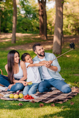 Happy family with one child taking selfie with smartphone while sitting on plaid at picnic