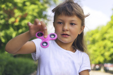 A small beautiful girl in a white T-shirt is disgusted and cautiously holding a pink spinner in her hand on the street.