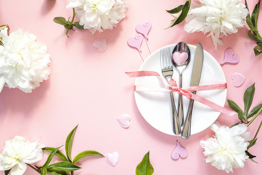 Festive table setting with cutlery, white peonies and hearts on pink table. Copy space.