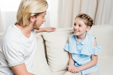 Fototapeta premium smiling father and daughter looking at each other and talking while sitting on sofa at home