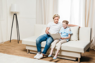 young father and daughter reading book while sitting on sofa at home