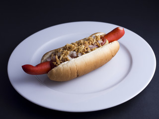Danish hot dog with red sausage, ketchup, mustard sauce and raw and fried onions on white plate, isolated on black background
