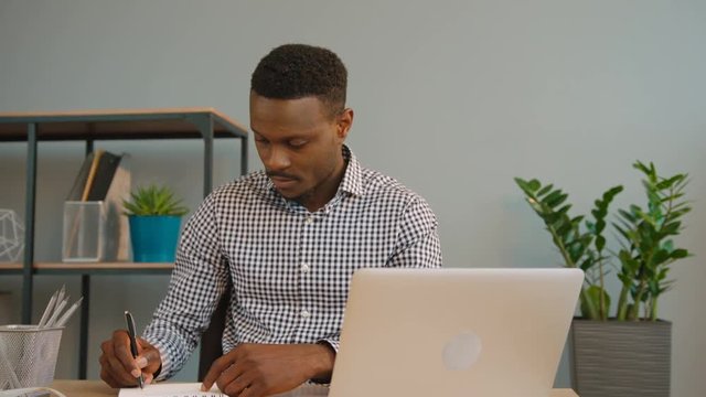 African americam business man working at the office. Black man writing down in the notebook from laptop computer