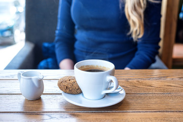 Coffee cup with cookie on wooden table in restaurant with view on window and plant. Woman on background.