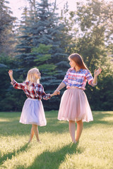 Naklejka premium Portrait of two smiling funny Caucasian girls sisters in plaid shirt and pink tutu tulle skirt, standing dancing barefoot on grass in park forest meadow at sunset. Friends having fun together.