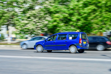 Fototapeta premium Car rides on city road with ghost effect. Buildings, green trees and parked cars on background.