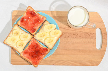 Square toast with jam and banana on a blue plate on a wooden board and a mug of milk